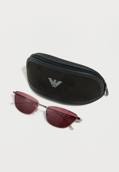 Ochelari de soare eleganți cu design cat-eye, lentile cu nuanță roz și rame subțiri aurii, așezați lângă o husă neagră cu fermoar și un logo argintiu, pe fundal alb.