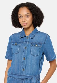 Combinaison en denim à manches courtes de couleur bleu clair, présentant un col, deux poches poitrine, des boutons en argent et un détail de taille ceinturée.