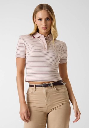 Kobieta z długimi blond włosami ma na sobie jasnoróżowy, pasiasty crop top polo i beżowe spodnie z ciemnobrązowym paskiem, stoi na tle jednolitego tła.