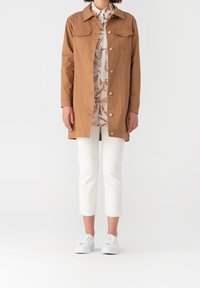 Giacca leggera marrone con colletto, chiusura a bottoni e due tasche sul petto. Indossata sopra una camicia a fiori e abbinata a pantaloni bianchi a 3/4 e sneakers.
