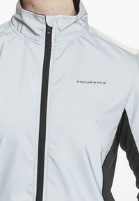 Lys grå atletisk jakke med høj krave, sorte detaljer og en frontlynt. Har ordet "ENDURANCE" på brystområdet.