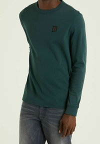 Långärmad, teal sweatshirt med ribbad textur och rund halsringning. Har en liten svart lapp med ett triangulärt mönster på bröstet.