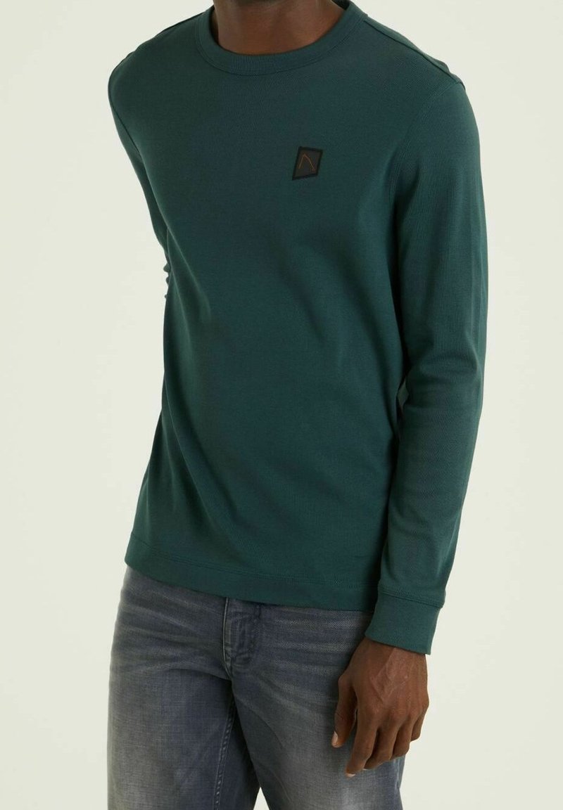 Långärmad, teal sweatshirt med ribbad textur och rund halsringning. Har en liten svart lapp med ett triangulärt mönster på bröstet.