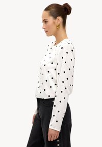 Blouse blanche à manches longues avec des pois noirs, col rond, texture lisse, associée à un pantalon foncé avec des boutons ornés sur le côté.