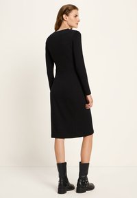 Eine Frau trägt ein enganliegendes schwarzes Maxikleid mit langen Ärmeln, kombiniert mit schwarzen Lederstiefeln bis zur Mitte der Wade, und steht vor einem schlichten hellen Hintergrund.