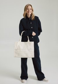 Canvas draagtas in crème met gegraveerde tekst "HEY MARLY HEY" over het midden, voorzien van twee stevige handvatten en eenvoudige ontwerpdetails.