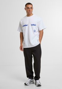 Hvid bomuld t-shirt med blå tekst "another cotton" og et rundt logo. Løs pasform med korte ærmer, parret med sorte bukser og sneakers.