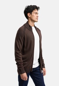 Brązowy kardigan zapinany na zamek z ribbowym kołnierzem i mankietami, z teksturowanym wzorem dzianiny, noszony na białej koszuli i w połączeniu z ciemnymi dżinsami.