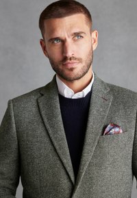 Blazer en chevrons gris avec revers en pointe, superposé sur un pull bleu marine. Un mouchoir de poche à motif coloré ajoute du détail. Le col de la chemise blanche est visible.