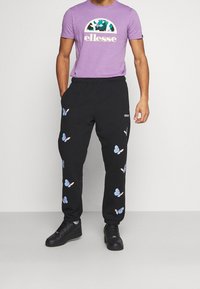 Lila t-shirt med en logotyp och svarta träningsbyxor med färgglada fjärilsmönster. Svarta sneakers kompletterar outfiten. Kanterna är elastiska.