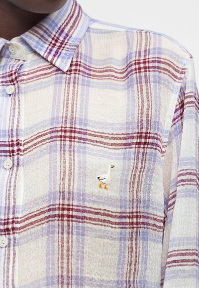 Κοντινή λήψη ενός λευκού, μωβ και κόκκινου καρό πουκαμίσου με λευκά κουμπιά και μια μικρή κεντημένη λευκή χήνα πάνω από την τσέπη στο στήθος.