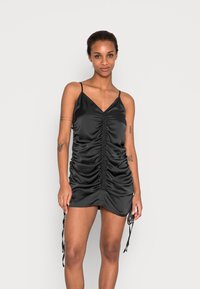 Vestido mini de satin preto com franzido lateral ajustável, decote em V, alças finas e laços decorativos na bainha. Textura suave, design ajustado.