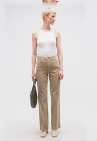 Άτομο με κοντά ξανθά μαλλιά που φοράει λευκό αμάνικο τοπ και μπεζ παντελόνι, κρατώντας μια γκρι τσάντα με υφή, στέκεται μπροστά σε λευκό φόντο.