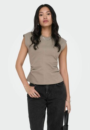 Jonge vrouw met lang zwart haar, gekleed in een mouwloos beige topje en zwarte spijkerbroek, die met één hand een zwarte handtas vasthoudt.