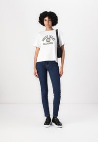 Bílá tričko s grafikou "Guess Jeans California", kombinované s tmavě modrými úzkými džíny a černými teniskami. Model drží černou crossbody tašku.