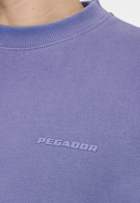 Primo piano di una persona che indossa una maglietta a girocollo viola con la scritta "PEGADOR" stampata in lettere in rilievo sul petto.