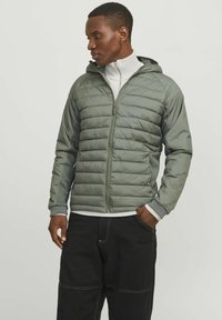 Grüne Puffjacke mit Kapuze, mit gesteppten Abschnitten, Reißverschluss vorne und gerippten Bündchen, getragen über einem weißen Oberteil mit hohem Kragen.