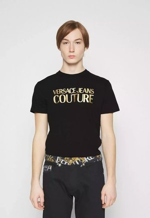 T-shirt nera in cotone con la scritta "VERSACE JEANS COUTURE" in lettere dorate. Abbinata a pantaloni neri e una cintura stampata. Design semplice e aderente.