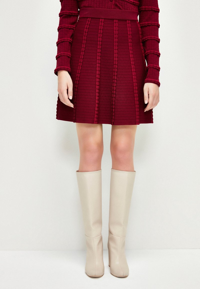 Jupe mini texturée bordeaux avec des côtes verticales, associée à des bottes beiges jusqu'aux genoux. La jupe possède une taille marquée et un ourlet évasé.