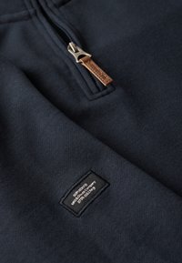 Sudadera de marine con media cremallera, diseño con herrajes plateados, tira de cuero y detalle de etiqueta cosida en el borde inferior. Textura suave.
