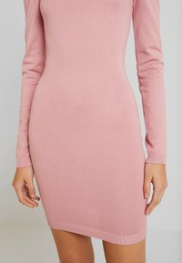 Femme portant une robe en maille rose ajustée, manches longues, longueur genou avec poignets et ourlet côtelés, montrant la moitié du corps du cou aux cuisses.