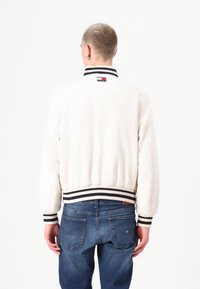 Hvit bomberjakke med svart-hvitt stripet ribbestrikket krage og mansjetter. Har en liten logo ved nakken. Brukt med blå jeans.