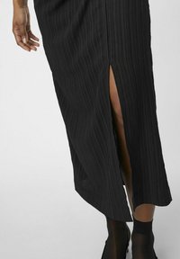 Robe midi noire côtelée avec une fente sur le côté, présentant un tissu texturé et un ourlet évasé. Le modèle porte des collants noirs transparents et des chaussures.