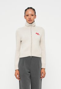 Pull en beige raccourci avec un col montant, fermeture éclair, poignets et ourlet côtelés, avec une broderie rouge "Alpha Club" sur la poitrine.