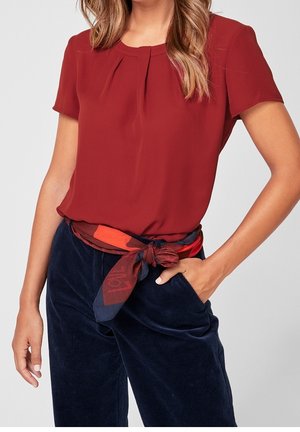 Mujer con blusa roja de manga corta metida dentro de pantalones de pana azul marino, con un cinturón de pañuelo rojo y azul con estampado, con la mano en el bolsillo.