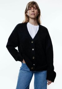 Schwarze Strickjacke mit V-Ausschnitt, fünf großen Knöpfen, lockerer Passform, gerippten Bündchen und einer weichen Textur, kombiniert mit hellblauen Jeans.