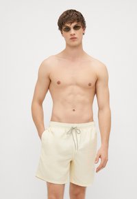 Bañadores de color amarillo claro con bolsillos laterales, cinturilla elástica y cordón gris. El modelo está de pie contra un fondo blanco, usando gafas de sol.