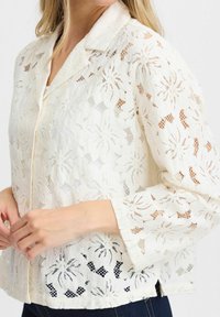 Vit spetsblus med blommönster, långa ärmar och krage. Utförliga texturer och transparenta sektioner framhäver den intrikata designen.