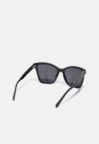 Ochelari de soare tip cat-eye cu rame din plastic negru, lentile de culoare gri închis și o textură elegantă, lucioasă. Prezintă accente de marcă pe laterale.