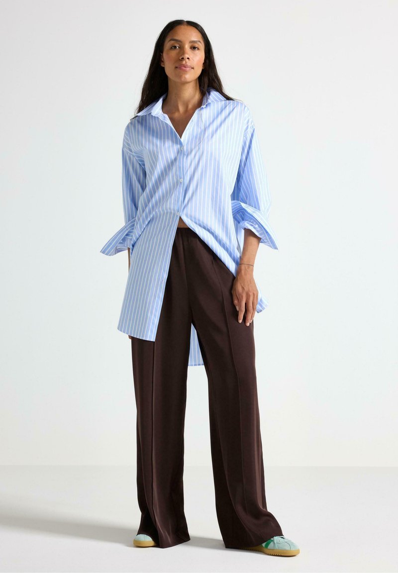 Camicia button-up a righe blu chiaro e bianche con maniche lunghe, abbinata a pantaloni larghi marroni e scarpe con dettagli verde menta.