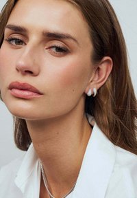 Silverörhängen med en texturerad, böjd design, kompletterat med en enkel vit blus och ett subtilt halsband. Naturlig sminkning framhäver ansiktsdrag.