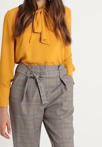 Gelbe Bluse mit Halsband, kombiniert mit karierten hochtaillierten Hosen mit einem Gürtel. Glattes Material mit einem karierter Muster in Grau und Braun.