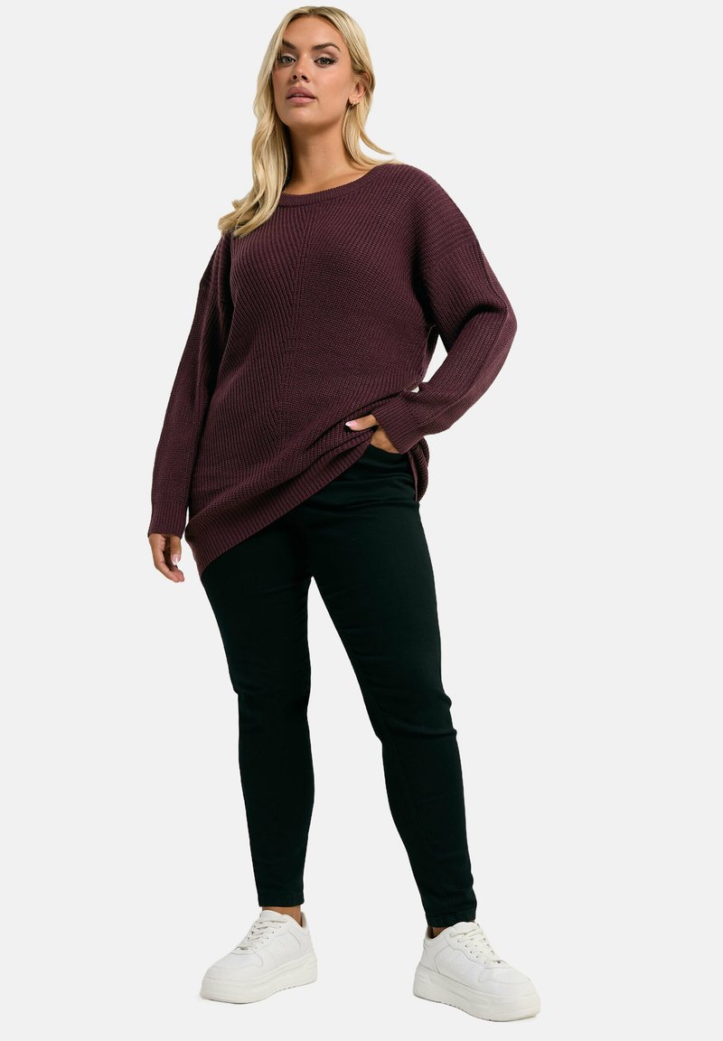 Femme portant un pull en tricot bordeaux ample, un pantalon noir skinny et des baskets blanches, se tenant avec une main dans la poche contre un fond uni.