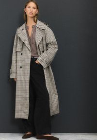 Beige und marineblauer, taillierter Karo-Trenchcoat mit doppelter Knopfleiste, großen Revers, langen Ärmeln und lockerer Passform. Getragen über einem grauen Oberteil und schwarzer Hose.