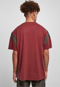 Camiseta de algodón color granate con mangas cortas, con acentos diagonales grises en los hombros y un corte holgado. Superficie texturizada, diseño sencillo.