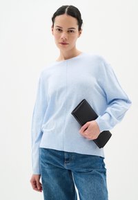 Vaaleansininen villapaita pitkillä hihoilla, pyöreällä kaula-aukolla ja hienovaraisella tekstuurilla; malli pitää mustaa clutch-laukkua. Farkut täydentävät asukokonaisuutta.