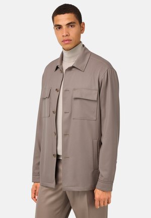 Chaqueta de tela gris mezclada con cuello, dos bolsillos en el pecho y cierre de botones. Lleva puesta una camiseta de cuello alto clara y unos chinos a juego.