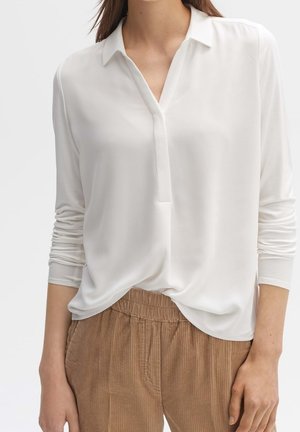 Femme portant un chemisier blanc à manches longues avec un col en V et un pantalon beige en velours côtelé avec une taille élastique.