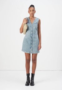 Denimkjole, lyseblå, V-hals, ermeløs, knappestolpe foran, A-linjeform. Modellen holder en beige veske, iført svarte ankelstøvler.