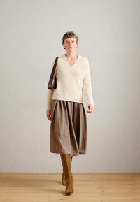 Κρέμα πλεκτό πουλόβερ με V-λαιμόκοψη, συνδυασμένο με μια καφέ πλισέ φούστα. Οι καφέ μπότες μέχρι το γόνατο και μια καφέ τσάντα ώμου ολοκληρώνουν την εμφάνιση.