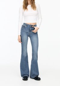 Jean flare bleu clair à taille haute, avec une conception classique à cinq poches et un léger effet délavé. Associé  à un haut blanc ajusté à manches longues.