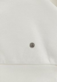Tessuto a maglia bianco con orlo a coste testurizzato e un badge rotondo in metallo con l'iscrizione "Key Largo".
