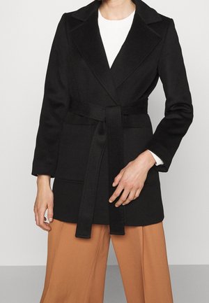 Persoon die een zwarte wollen jas met riem draagt over een wit shirt en beige wijde broek, tegen een effen grijze achtergrond.