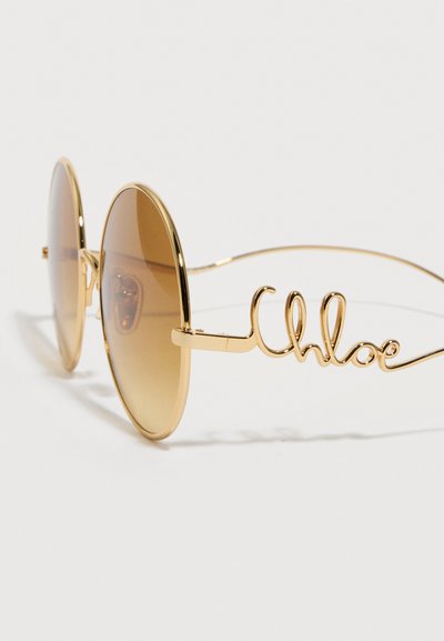 Στρογγυλά γυαλιά ηλίου με καφέ σκουρόχρωμους φακούς και χρυσά σύρματα στους βραχίονες που σχηματίζουν τη λέξη "Chloe" στο πλάι.