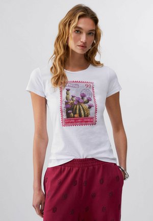 Giovane donna con capelli biondi ondulati che indossa una t-shirt bianca con un design colorato di cactus e pantaloni rossi con motivi neri.
