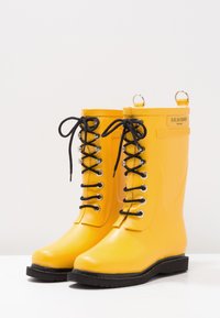 Bottes de pluie en caoutchouc jaune avec des lacets noirs, des œillets en métal et des languettes. Texture lisse et bout arrondi. Semelle noire avec une légère crantage.